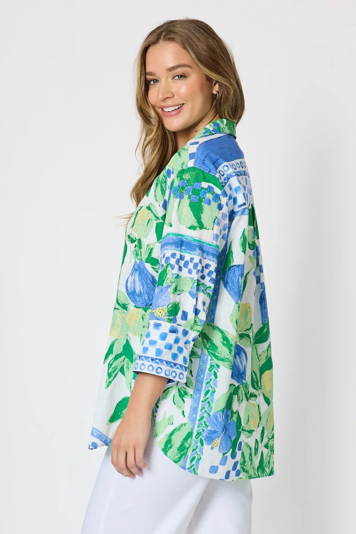 Woman wearing a button up shirt with collar and Mediterranean print with botanical, lemon and blue geometric design and white pants and white background.