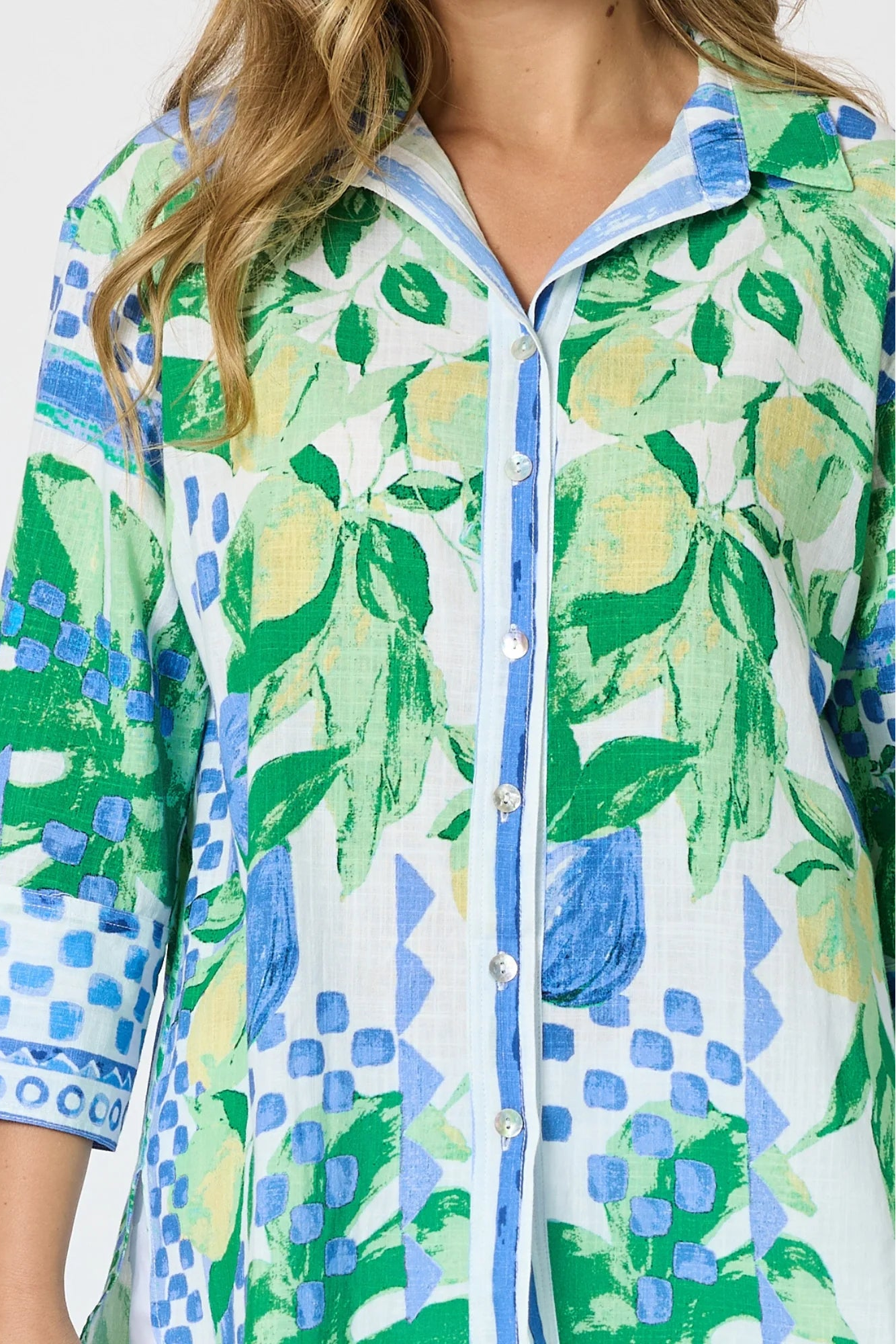 Woman wearing a button up shirt with collar and Mediterranean print with botanical, lemon and blue geometric design and white pants and white background.