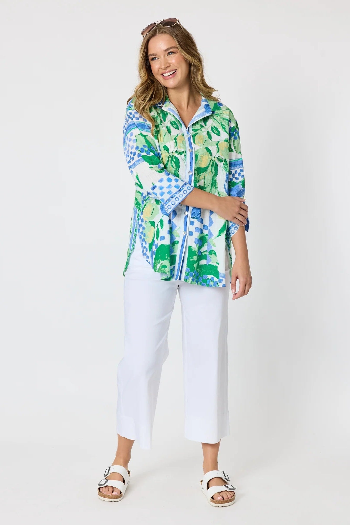 Woman wearing a button up shirt with collar and Mediterranean print with botanical, lemon and blue geometric design and white pants and white background.