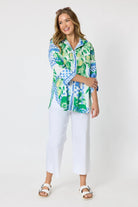 Woman wearing a button up shirt with collar and Mediterranean print with botanical, lemon and blue geometric design and white pants and white background.