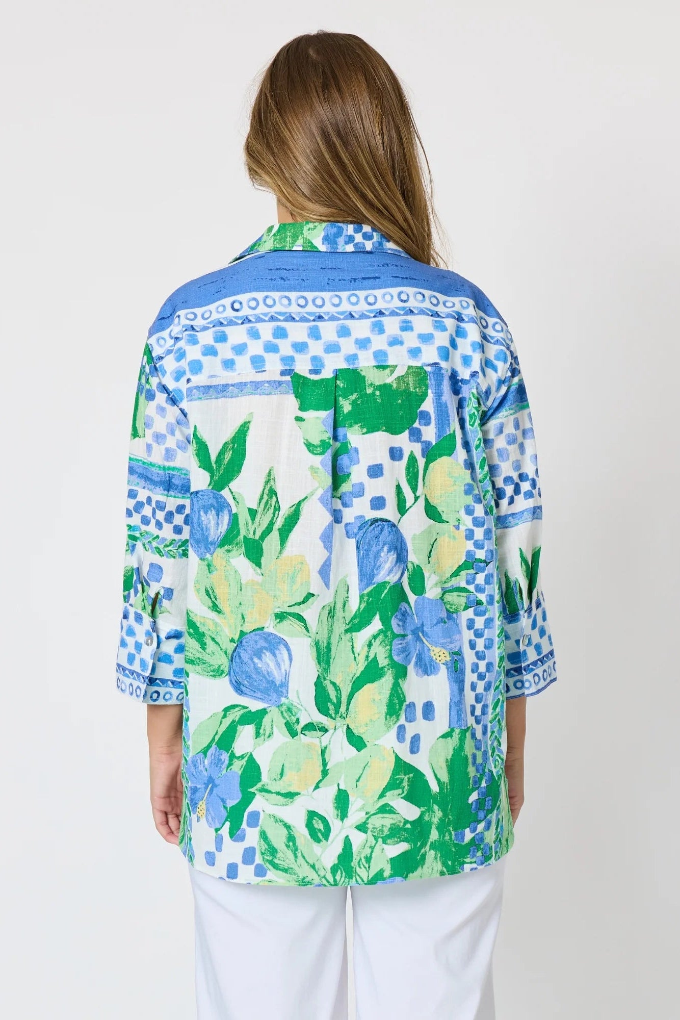 Woman wearing a button up shirt with collar and Mediterranean print with botanical, lemon and blue geometric design and white pants and white background.