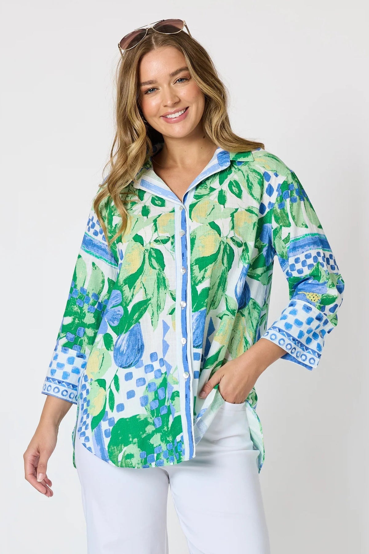 Woman wearing a button up shirt with collar and Mediterranean print with botanical, lemon and blue geometric design and white pants and white background.