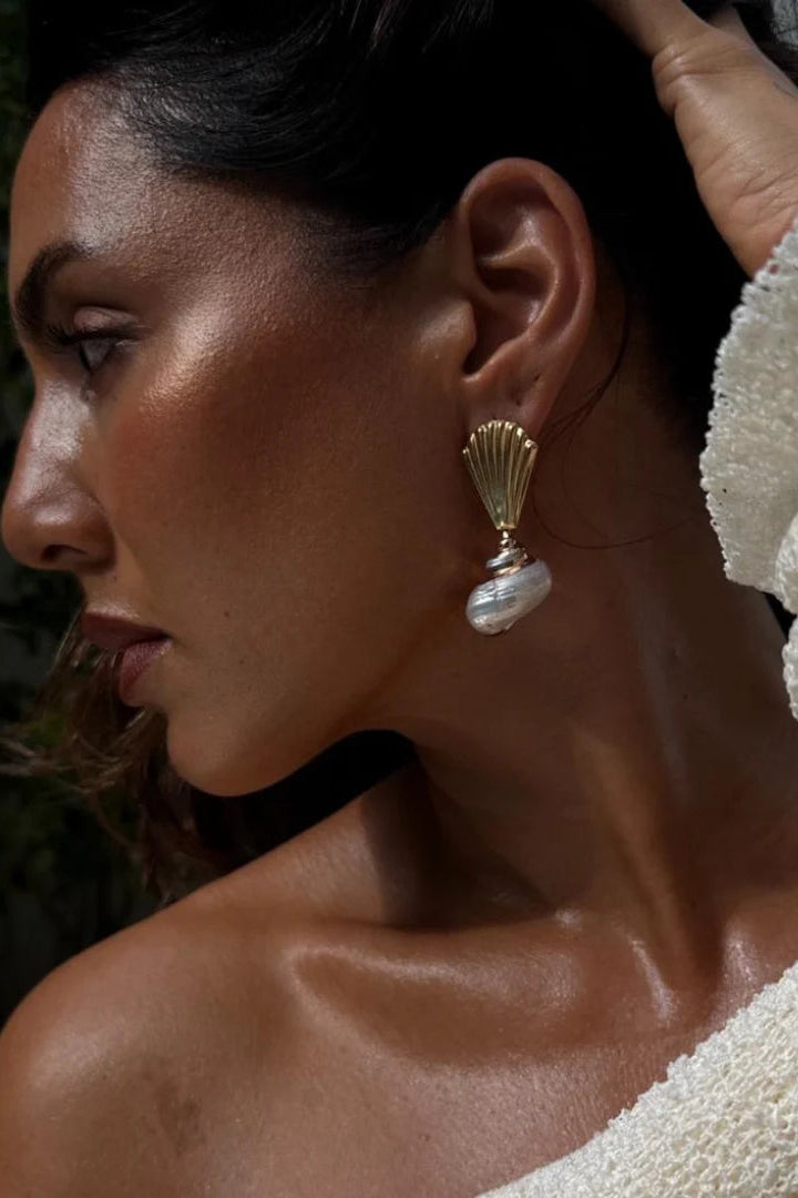 A close-up side profile of a woman wearing a gold-toned dangle earring with a white pearl-like drop, paired with a textured cream off-the-shoulder top.