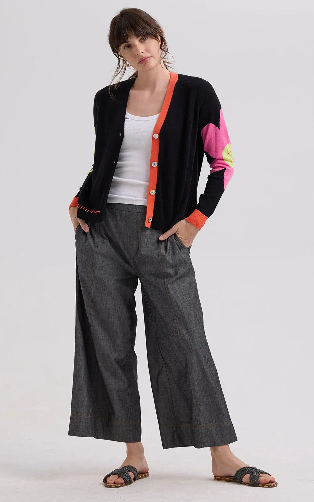 Woman wearing a black cardigan with colorful sleeves, white tank top, gray wide-leg pants, and black sandals on a white background.