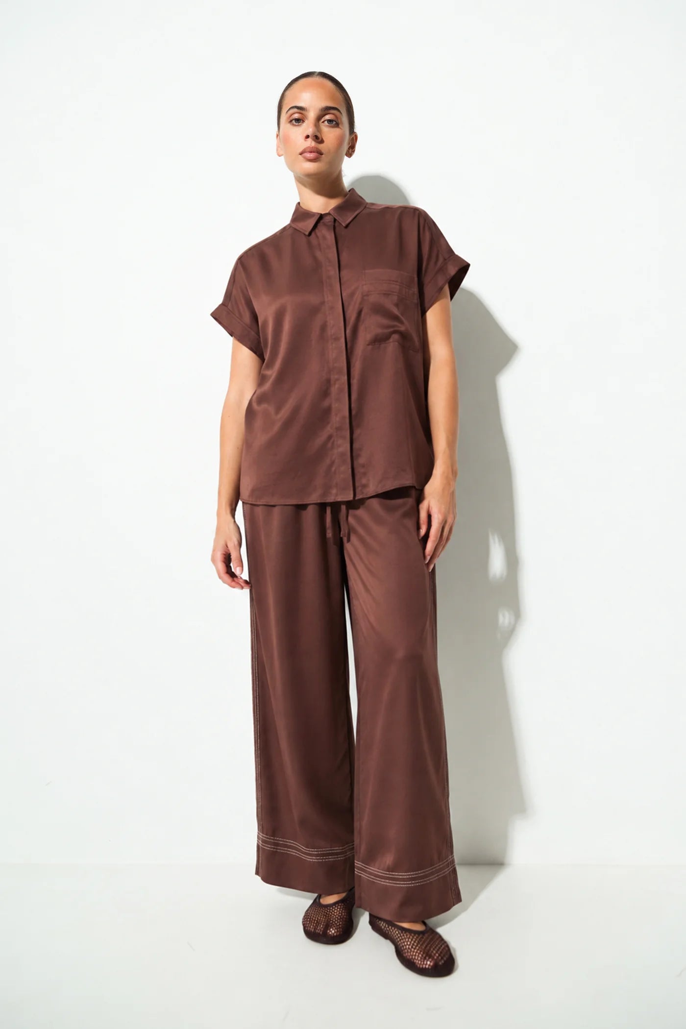 A woman stands against a plain white studio wall wearing a matching brown satin-like two-piece outfit with a short-sleeve collared top and wide-leg pants.