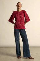 Woman wearing a red blouse with floral patterns and blue jeans against a plain background