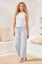 Woman wearing a white sleeveless top and light blue jeans standing in a room with beige cushions and a plant.