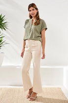Woman wearing a green top and beige pants standing in a bright room with plants.