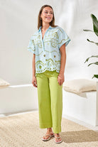 Woman wearing a light blue patterned top and green pants in a bright room.